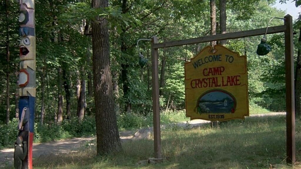 in a screenshot from the movie Friday the 13th, a sign reading Welcome to Camp Crystal Lake hangs on a simple wooden frame next to a native american totem pole