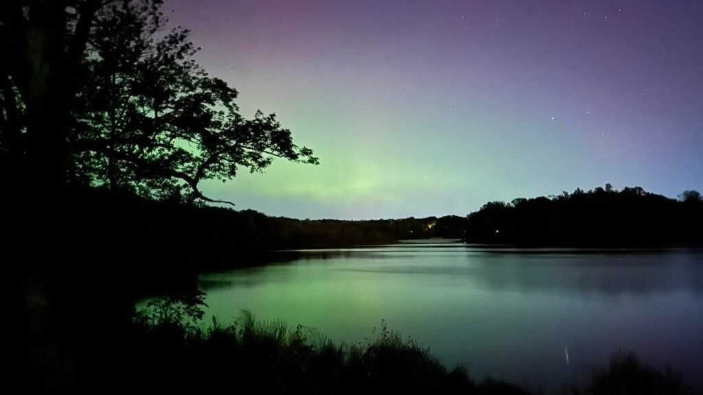 The Aurora Borealis Is Back: These 18 States Have a Chance to See It Over 2 Nights Green aurora reflected in a body of water with trees along the side.