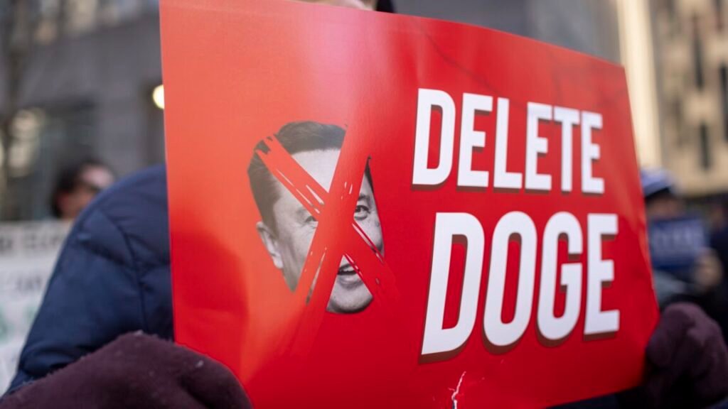 A protestor holds a sign that has a picture of Elon Musk with an X over his face, and text that says "Delete DOGE"