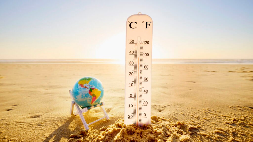 a sandy beach with a globe balanced in a beach chair next to an oversized thermometer showing high temperatures.