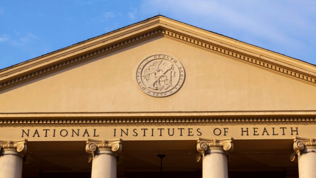 A view of the exterior of a building with a triangular top supported by pillars. Beneath a circular logo are the words "national Institutes of Health"