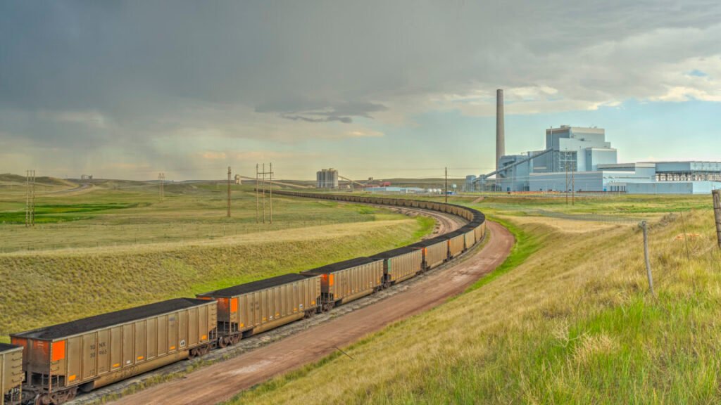 A grass covered prairie with rail line curving around to the left. The rails are filled with train cars carrying coal, and a large facility with a smokestack sits on the horizon.
