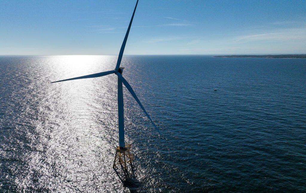 A aerial view of a large, sunlit expanse of ocean with a single wind turbine in it.