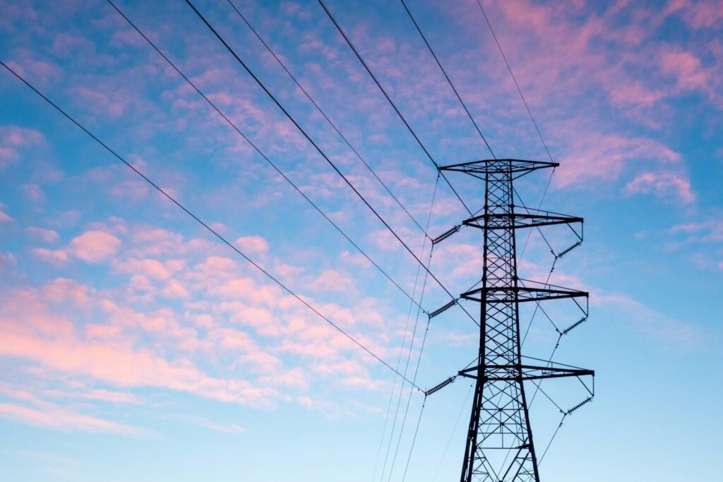 Trump Declared a Power Grid Emergency. The Parts It Restricts Already Have a 128 Week Wait. High voltage electricity transmission pylon and power lines silhouetted against an orange sky