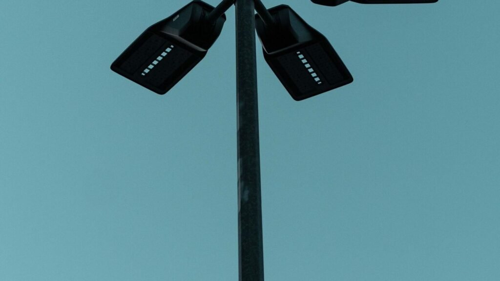 A $75 Shirt Makes You Invisible to AI Cameras. It Makes You Extremely Visible to People. A surveillance camera mounted on a streetlight against a blue sky, illustrating the AI object detection systems the Digital Camouflage shirt is designed to confuse