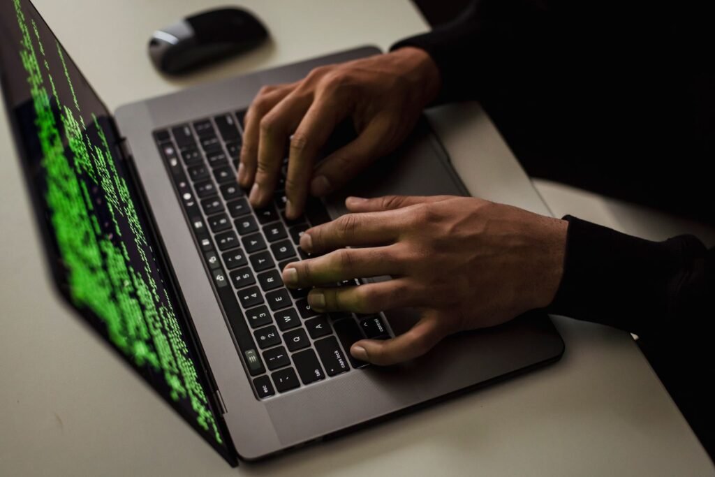 Close-up of hands typing on a laptop with green code on the screen, illustrating OAuth consent phishing and unauthorized email account access