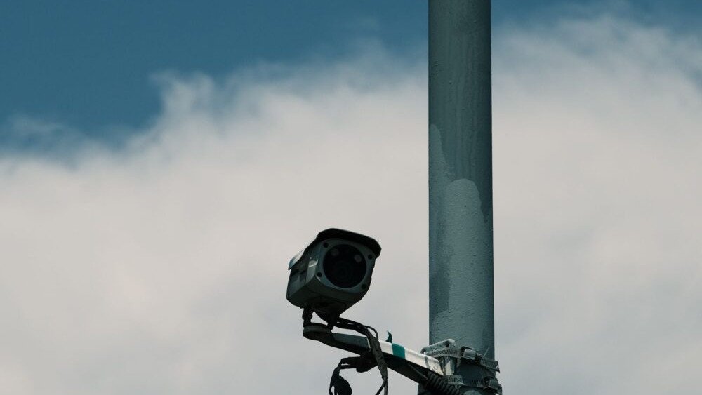 Hackers Pulled a Flock Camera Off a Pole. The Encryption Key Was Sitting Inside It. A traffic surveillance camera mounted on a metal pole against a cloudy sky, representing automated license plate reader hardware