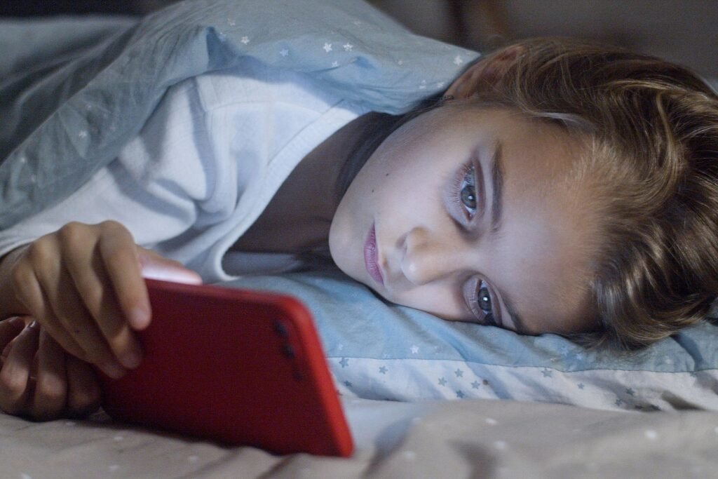 A child lying in bed under a blanket looking at a smartphone screen at night