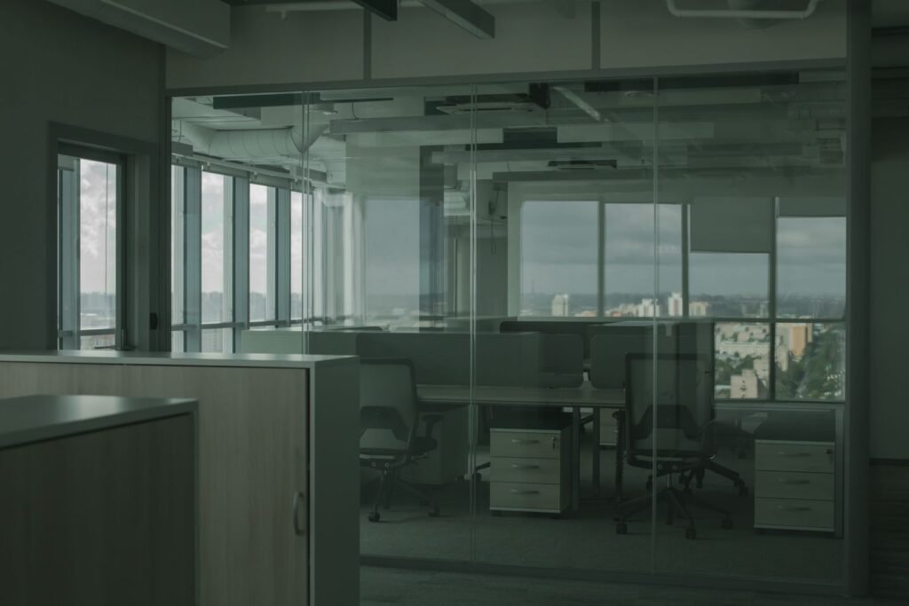 An empty open plan office with black rolling chairs pushed up to vacant desks beside glass walls