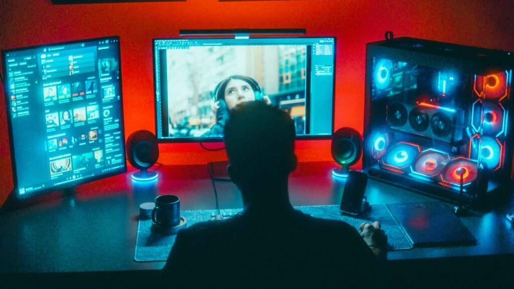Gamer seen from behind at a desk with two monitors and an RGB lit gaming PC in a red lit room