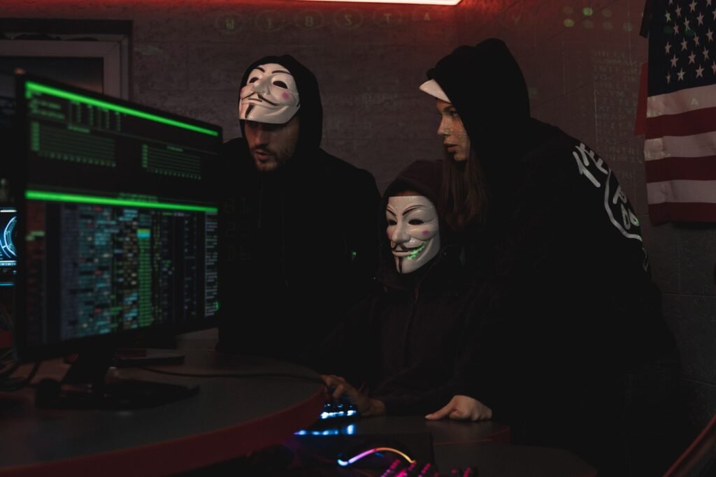 Three people in hoodies and masks gathered around glowing computer monitors in a dark room