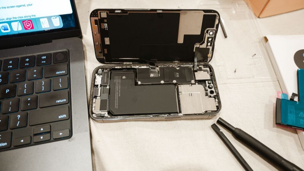 Opened iPhone on a work mat with its battery and logic board exposed, next to a laptop and plastic pry tools