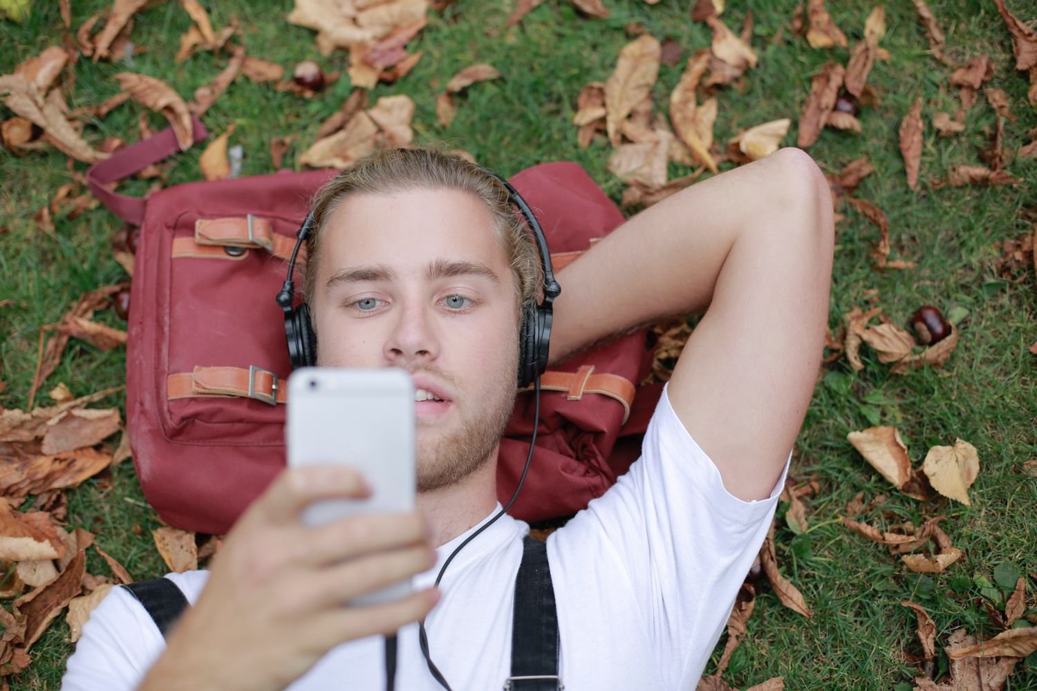 Young man lying on autumn leaves with headphones on, picking a song on his phone