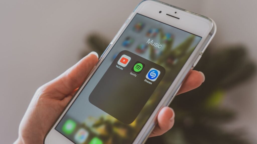 Hand holding an iPhone open to a folder of music apps including YouTube Music, Spotify and Shazam