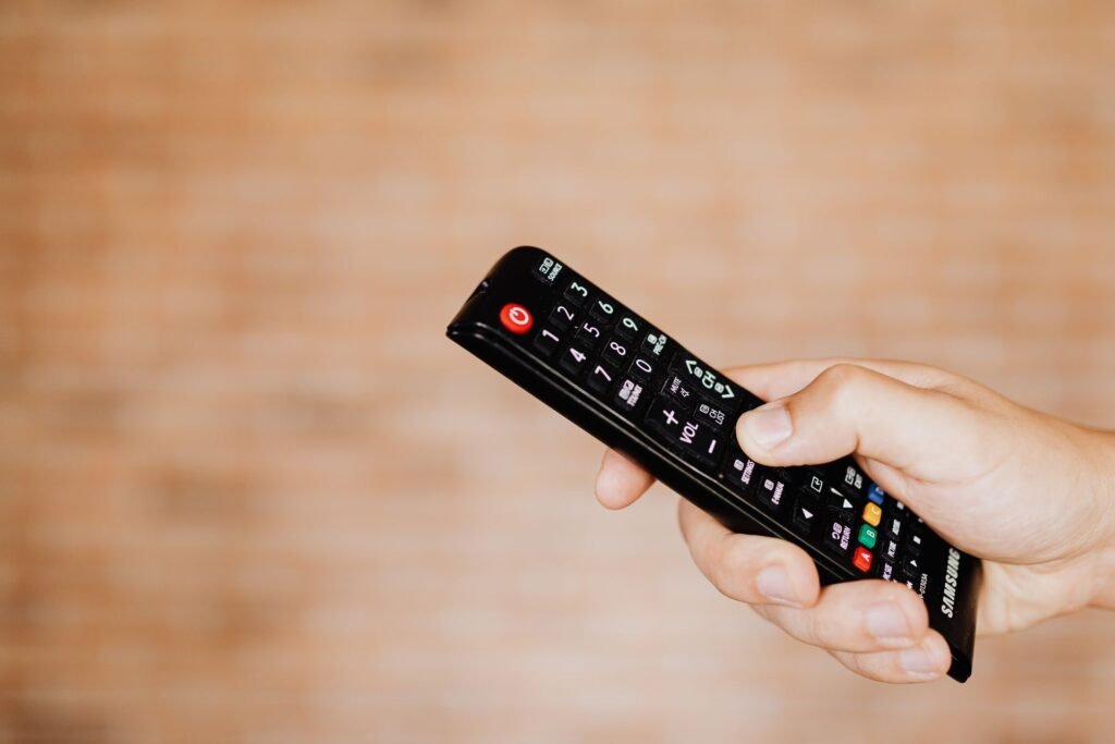 Hand holding a black TV remote control in front of a blurred brick wall